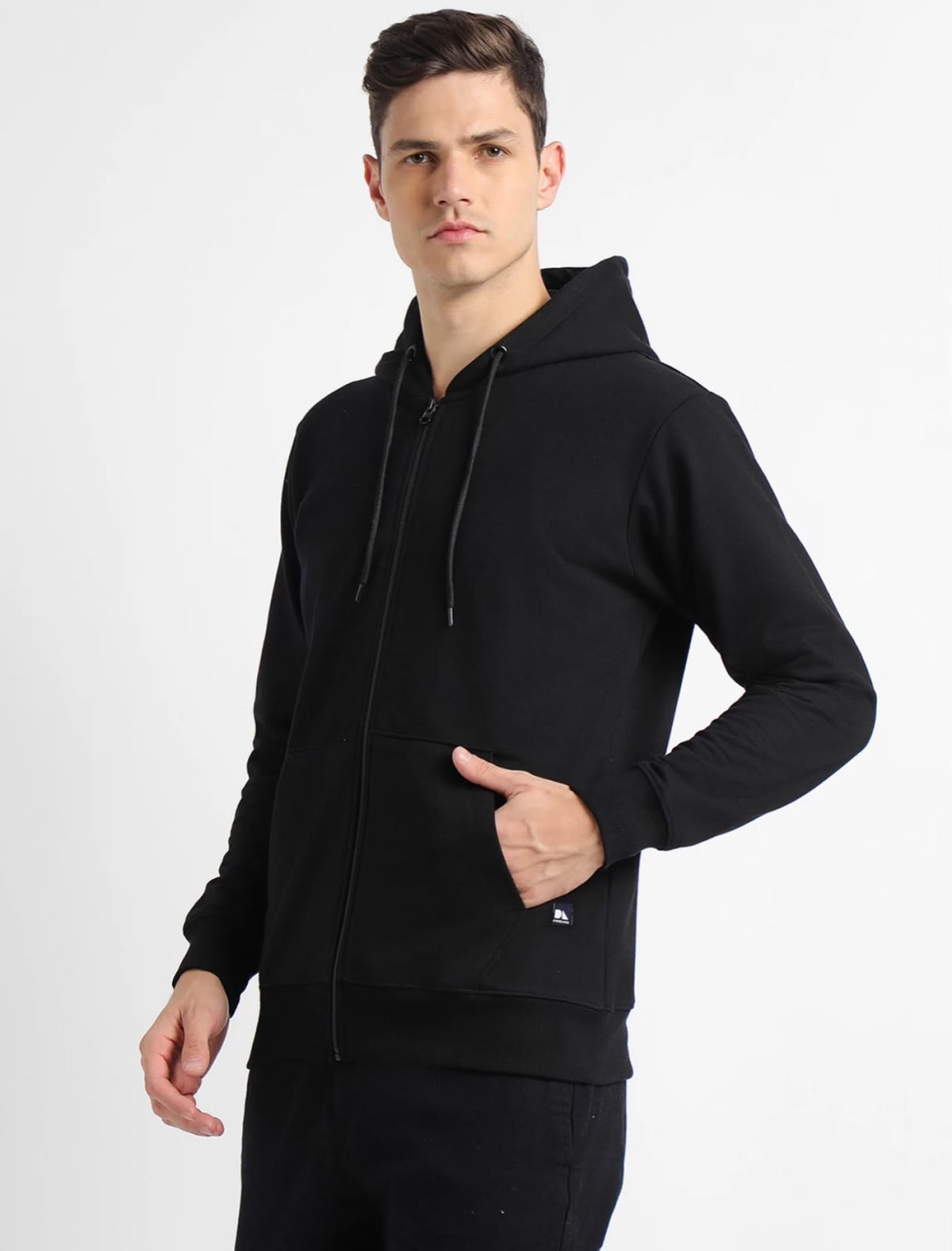 A man wearing a black full-zip hoodie with a drawstring hood and front kangaroo pocket, standing against a light gray studio background